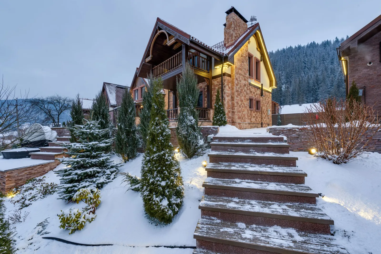 A large mountain house lit warmly at dusk with snow-covered grounds